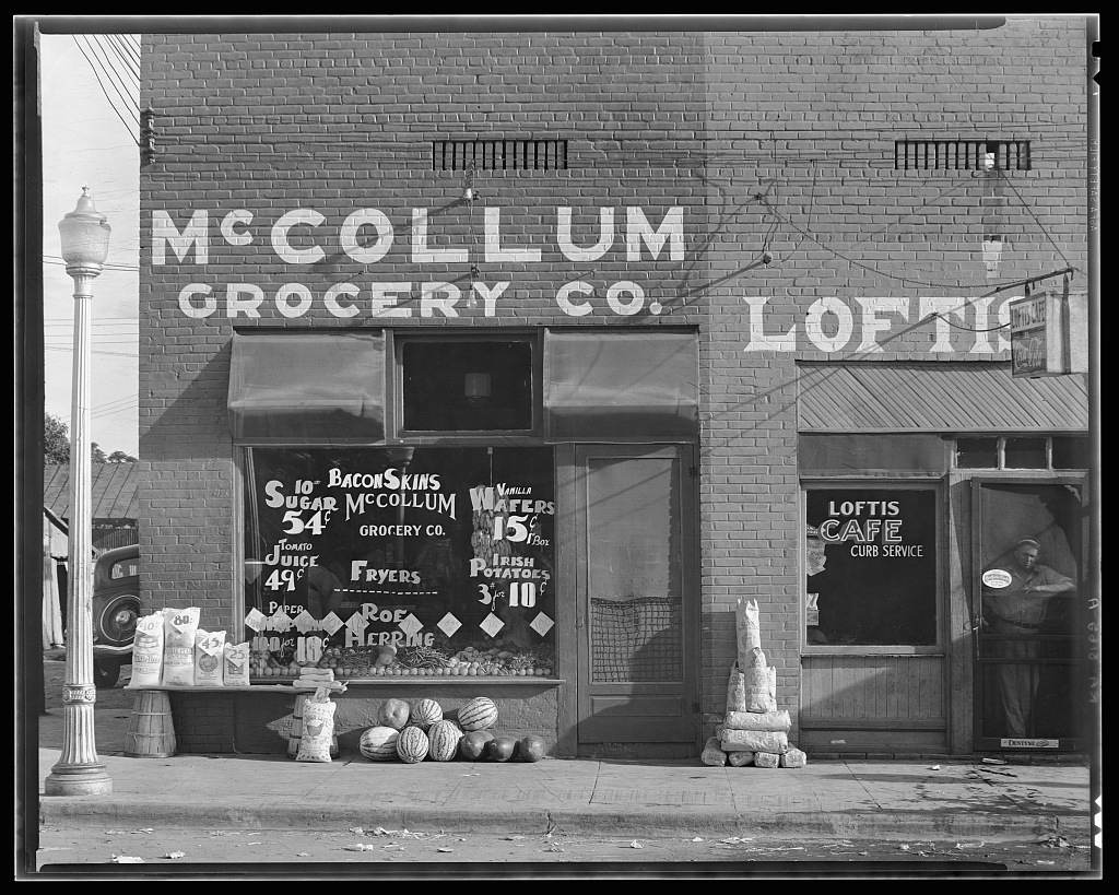 A grocery storefront in Greensboro, Alabama, photographed in 1936 for the Farm Security Administration: the windows are covered edge to edge in hand-painted signs quoting that week's prices by the pound.